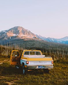 an old yellow truck parked in front of a mountain with snow capped mountains behind it