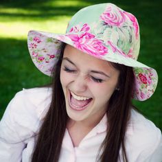 a young woman wearing a pink and green floral hat smiles at her cell phone while sitting in the grass