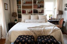 a bed with white sheets and pillows in a bedroom next to a window filled with books