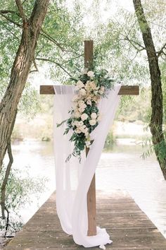 a cross decorated with white flowers and greenery on a wooden platform next to water