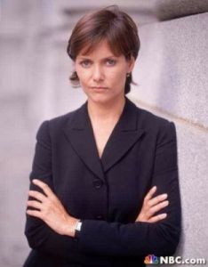 a woman with her arms crossed standing in front of a wall and looking at the camera