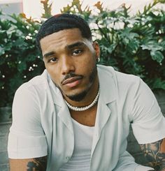 a man with tattoos sitting on the ground in front of some bushes and plants, wearing a white shirt