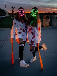 two people with neon masks on their faces and holding baseball bats in the street at sunset