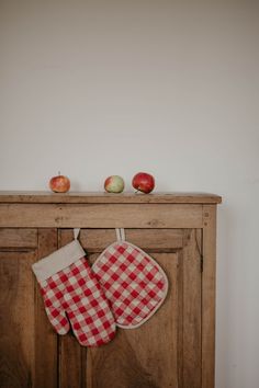 two red and white checkered oven mitts hanging from an old wooden cabinet with apples on top