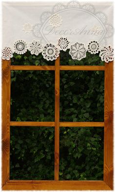 an open window with white lace on the top and green plants behind it in front of a wooden frame