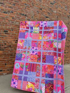 a pink and purple quilt sitting on the side of a brick wall next to a sidewalk