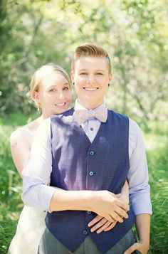 a young man and woman standing next to each other in front of green grass with trees behind them