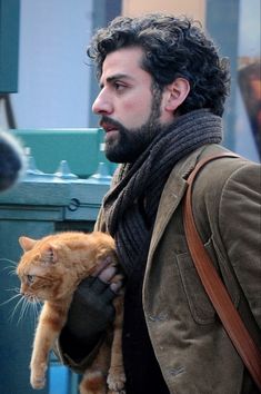 a bearded man holding an orange cat in his right hand while walking down the street