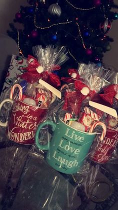 three coffee mugs with candy canes wrapped in cellophane are sitting next to a christmas tree