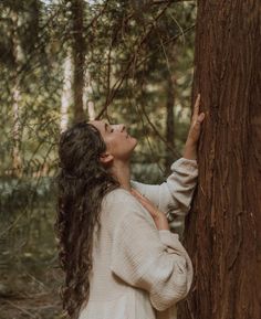 a woman standing next to a tree in the forest looking up at something on it