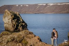 a man and woman standing next to each other near the water with mountains in the background