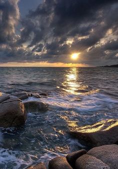 the sun is setting over the ocean with rocks in the foreground and clouds in the background