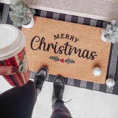 a person standing in front of a merry christmas door mat holding a cup of coffee