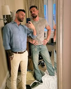 two men standing in front of a mirror looking at their cell phones and taking a selfie