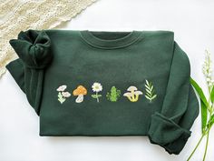 a green sweatshirt with embroidered mushrooms and flowers on the front, sitting next to a white lace doily