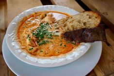 a white plate topped with soup and meat on top of a wooden table next to bread