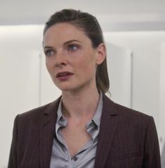 a woman standing in front of a white wall wearing a brown jacket and gray shirt