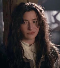 a woman with long hair standing in front of a wooden pole and looking at the camera