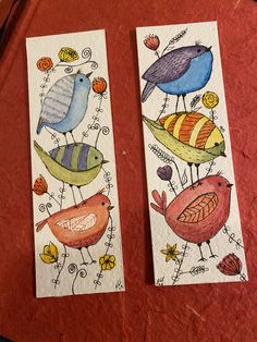 two bookmarks with birds painted on them sitting on a red table top next to each other