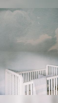 two cribs in a room with clouds painted on the wall and one is white