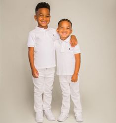 two young boys standing next to each other wearing white shirts and pants with their arms around each other