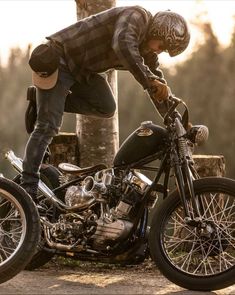 a man in plaid shirt and jeans leaning over a motorcycle with his hands on the wheel