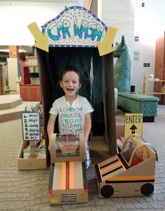 a young boy sitting in a cardboard car wash booth with boxes and signs on the floor