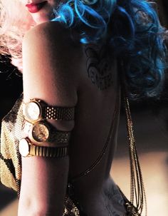 a woman with blue hair and tattoos on her arm wearing gold bracelets, holding a purse