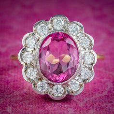 A spectacular Edwardian inspired daisy cluster ring bezel set with a vibrant oval cut pink topaz in the centre (approx. 5ct), haloed by twelve sparkling brilliant cut cubic zirconia petals. Pink topaz comes in shades of soft pink and fiery red and like a ruby is considered a stone of the heart. A symbol of femininity and romance, it's said to bring good fortune in love, providing happiness and vitality to any relationship. The ring is crafted in silver and the band is gilded in 18ct yellow gold. Heirloom Pink Ring With Halo Setting, Pink Cluster Ring Fine Jewelry, Classic Pink Diamond Cluster Ring, Exquisite Pink Ring With Halo Setting, Pink Cluster Ring With Gemstone, Pink Oval Cluster Ring Fine Jewelry, Exquisite Pink Halo Setting Ring, Pink Cluster Gemstone Ring, Pink Oval Halo Ring For Formal Occasions