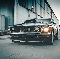 an old mustang driving down the road in front of a truck with its headlights on