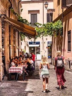 two women walking down an alley way with people sitting at tables in the back ground