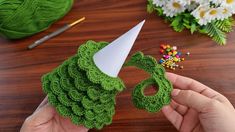 a hand holding a green crocheted hat on top of a wooden table next to flowers