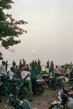 a group of people standing next to motorcycles on a dirt field near the ocean and trees