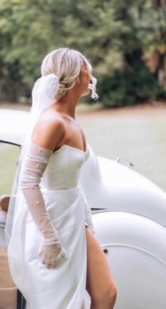 a woman in a white wedding dress and veil walking towards a car with her leg on the hood