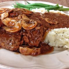 a white plate topped with meat covered in gravy next to mashed potatoes and green beans