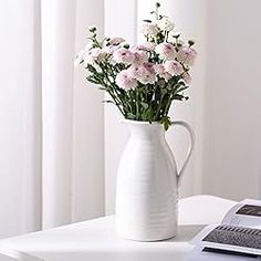 a white vase filled with pink flowers sitting on top of a table next to a window