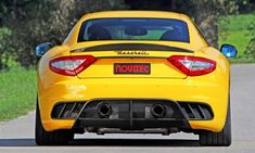 the rear end of a yellow sports car driving on a road with trees in the background