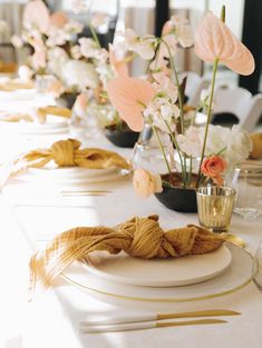 the table is set with white plates and gold place settings, along with flowers in vases