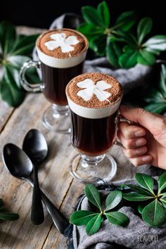 two people holding mugs filled with hot chocolate and topped with shamrock leaves, on top of a wooden table