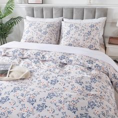 a white bed with blue and orange flowers on the comforter next to a potted plant