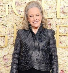 an older woman standing in front of a wall with roses on it and wearing a black jacket