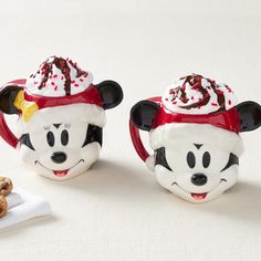 two mickey mouse mugs sitting next to each other on a white table with a cookie in front of them