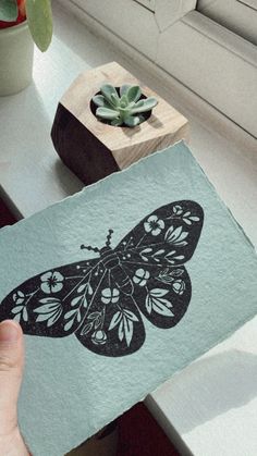 a person holding up a piece of paper with a butterfly on it next to a plant