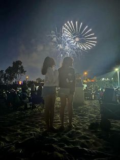 two women standing on top of a sandy beach next to a firework in the sky