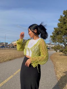 a woman standing on the side of a road wearing a green knitted sweater and black skirt