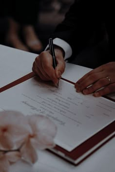 a person writing on a piece of paper with a pen in their hand and flowers around them