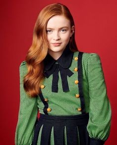 a woman with long red hair wearing a green and black dress standing in front of a red background