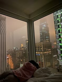 a person laying on a bed in front of two large windows with city lights behind them