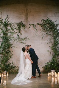 a bride and groom kissing in front of candles at their wedding ceremony with greenery on the wall behind them