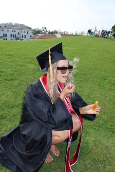 a woman in graduation gown sitting on the grass eating something out of her hand while wearing sunglasses